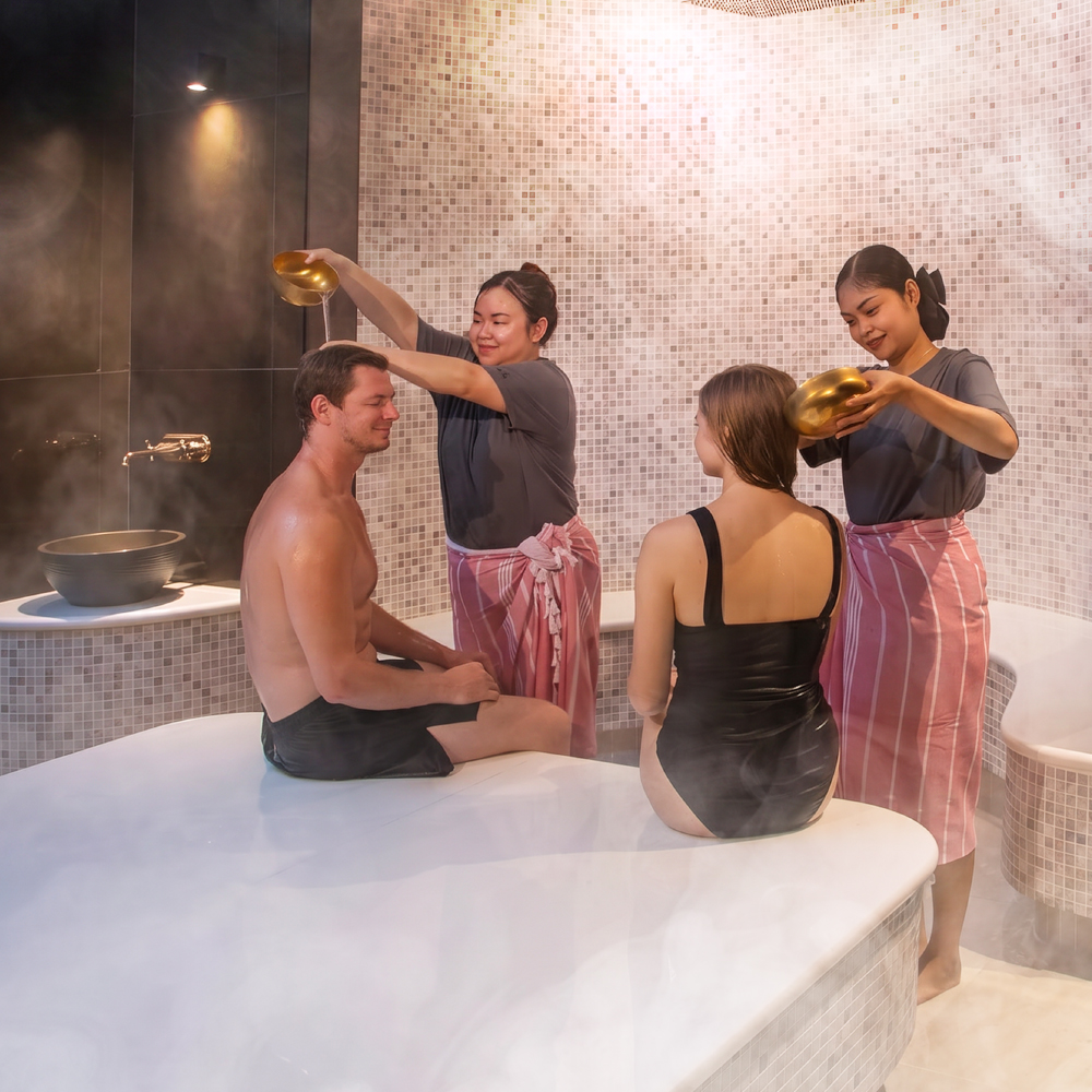 two happy customers sitting on a marble bed in the Thai Hammam room at Amla Aqua - Organic spa & hydrotherapy while two massage therapists are standing infront of them and pouring warm water on their heads to clean them after their Thai Hammam ritual